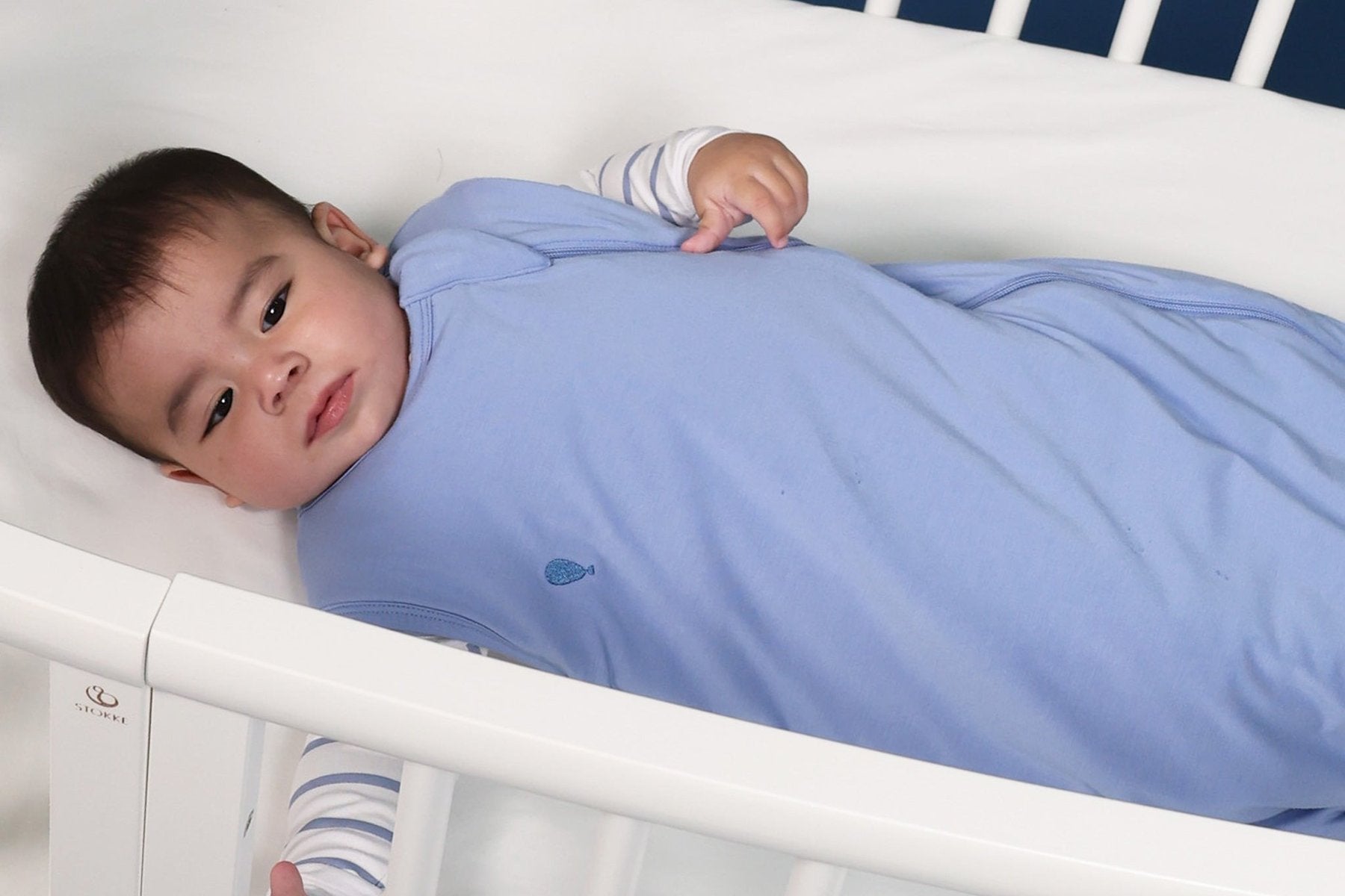 Baby lying on their back in a cot in the Atlas & Maya 1.0 TOG bamboo sleeping bag in Frost Blue