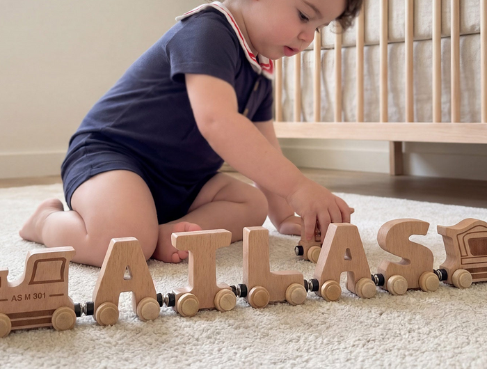 Child playing with a wooden toy train set spelling 'ATLAS' on a carpeted floor.